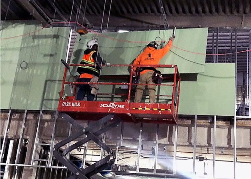 two workers on a lift are moving green wallboard into place on steel frames on an indoor construction site