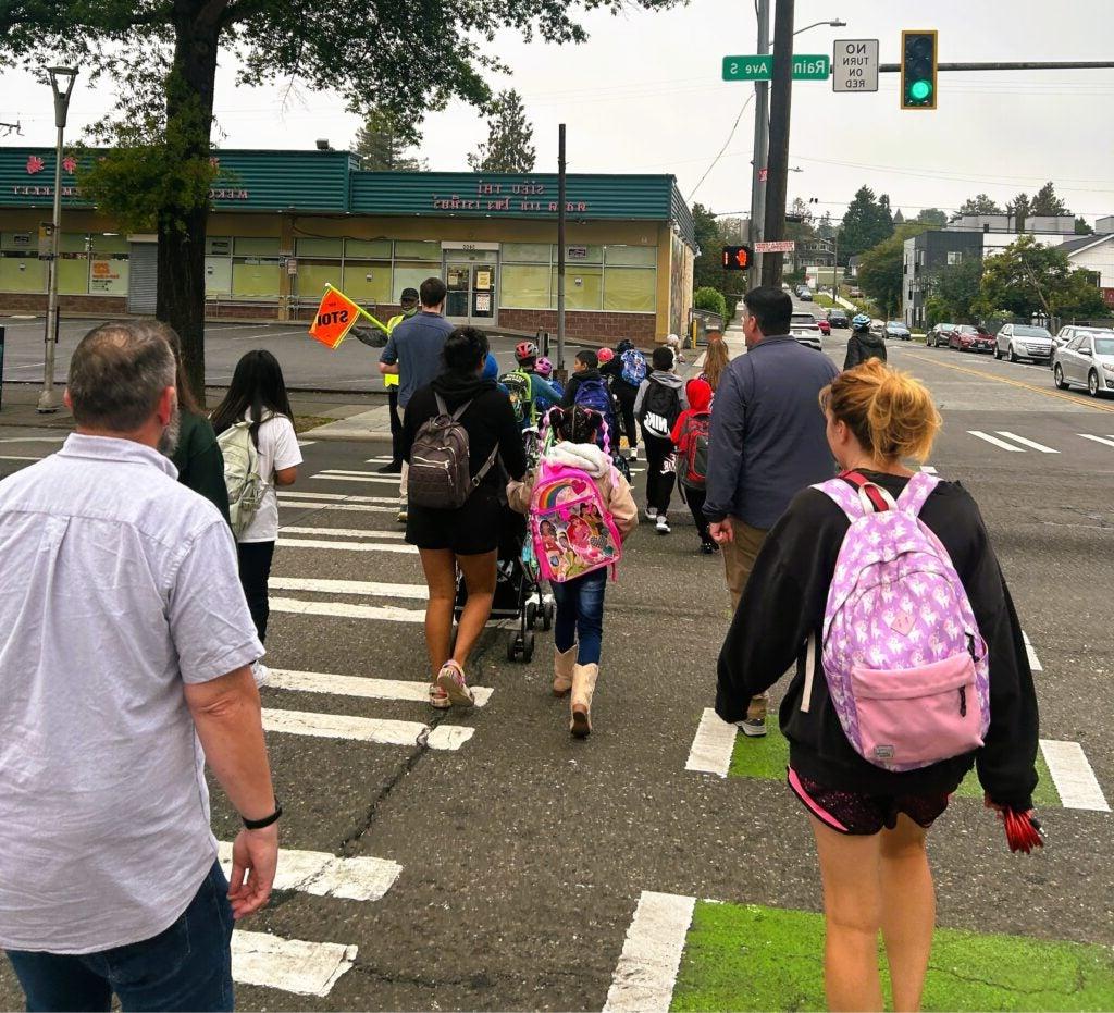 Students and families walk across Rainier Ave together with help of crossing guard.